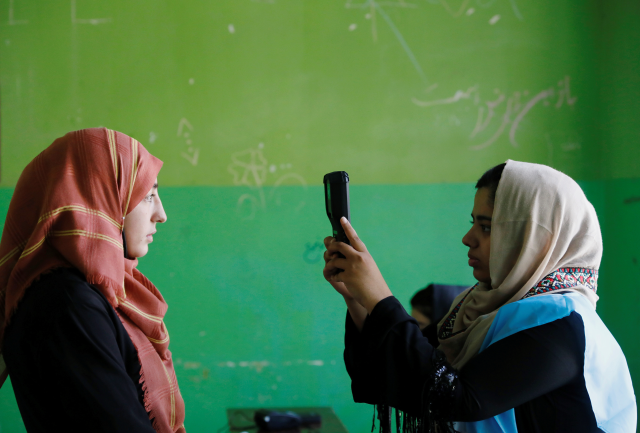 A woman holds up a device to take a picture of someone standing in front of a green wall